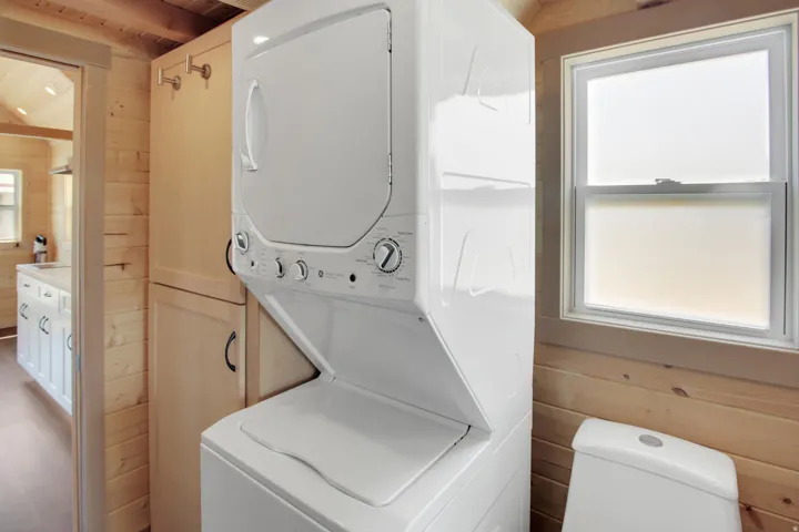Stackable washer and dryer unit, wood-paneled walls, built-in storage cabinet, white single-hung window, and a white toilet