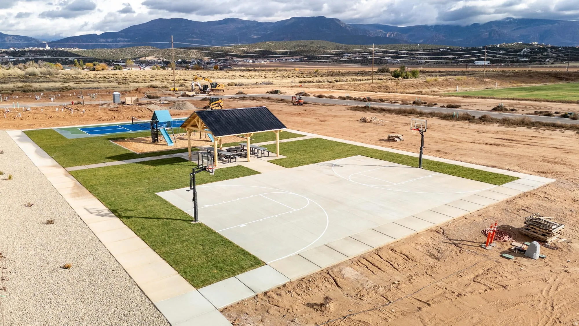 Recreational area featuring a concrete basketball court with painted lines, a covered picnic pavilion with tables, a blue playground structure, and a secondary sports court with blue surfacing