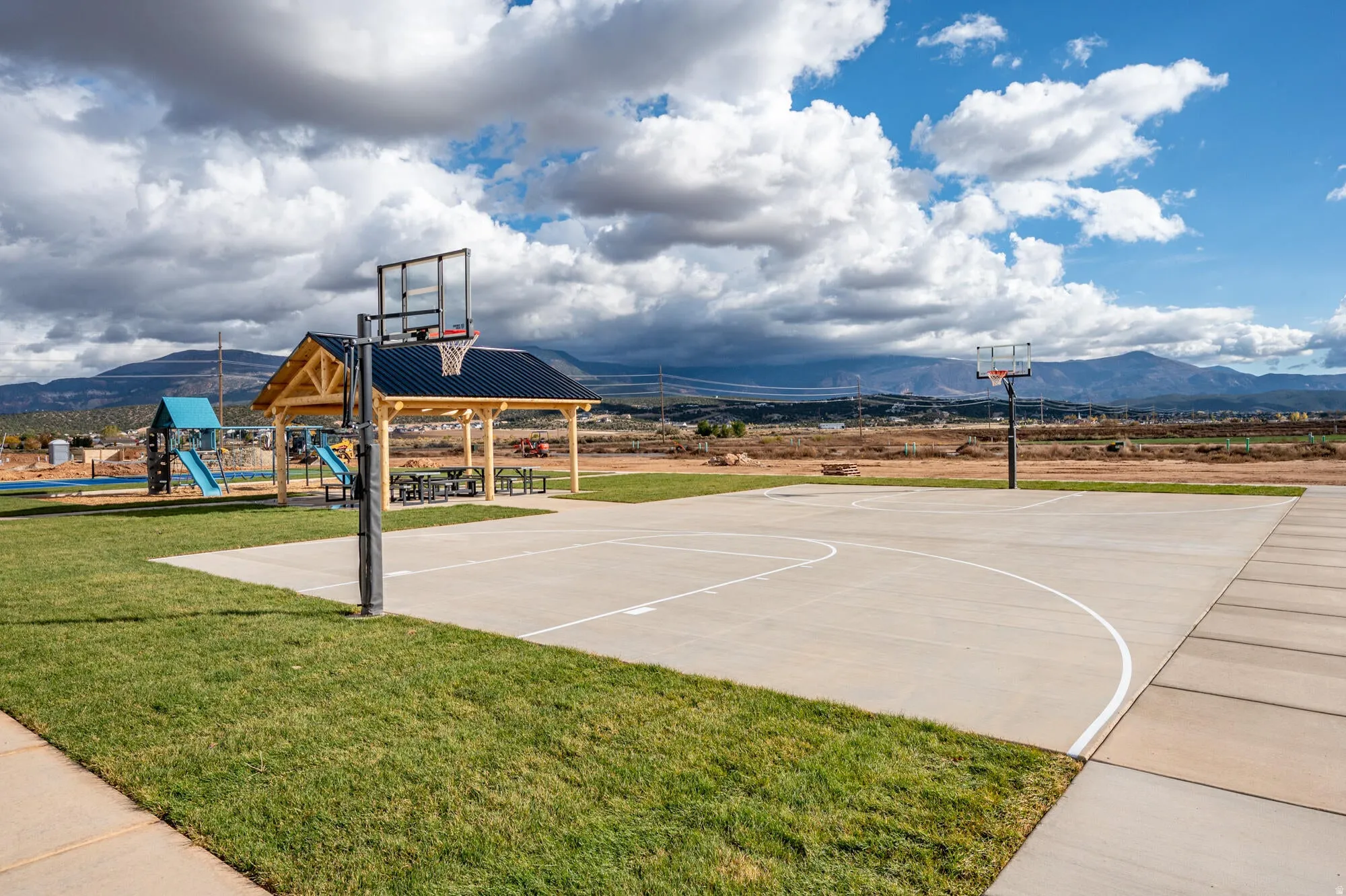 Concrete basketball court featuring two hoops, a covered picnic area with multiple tables, a playground with slides, and expansive green spaces