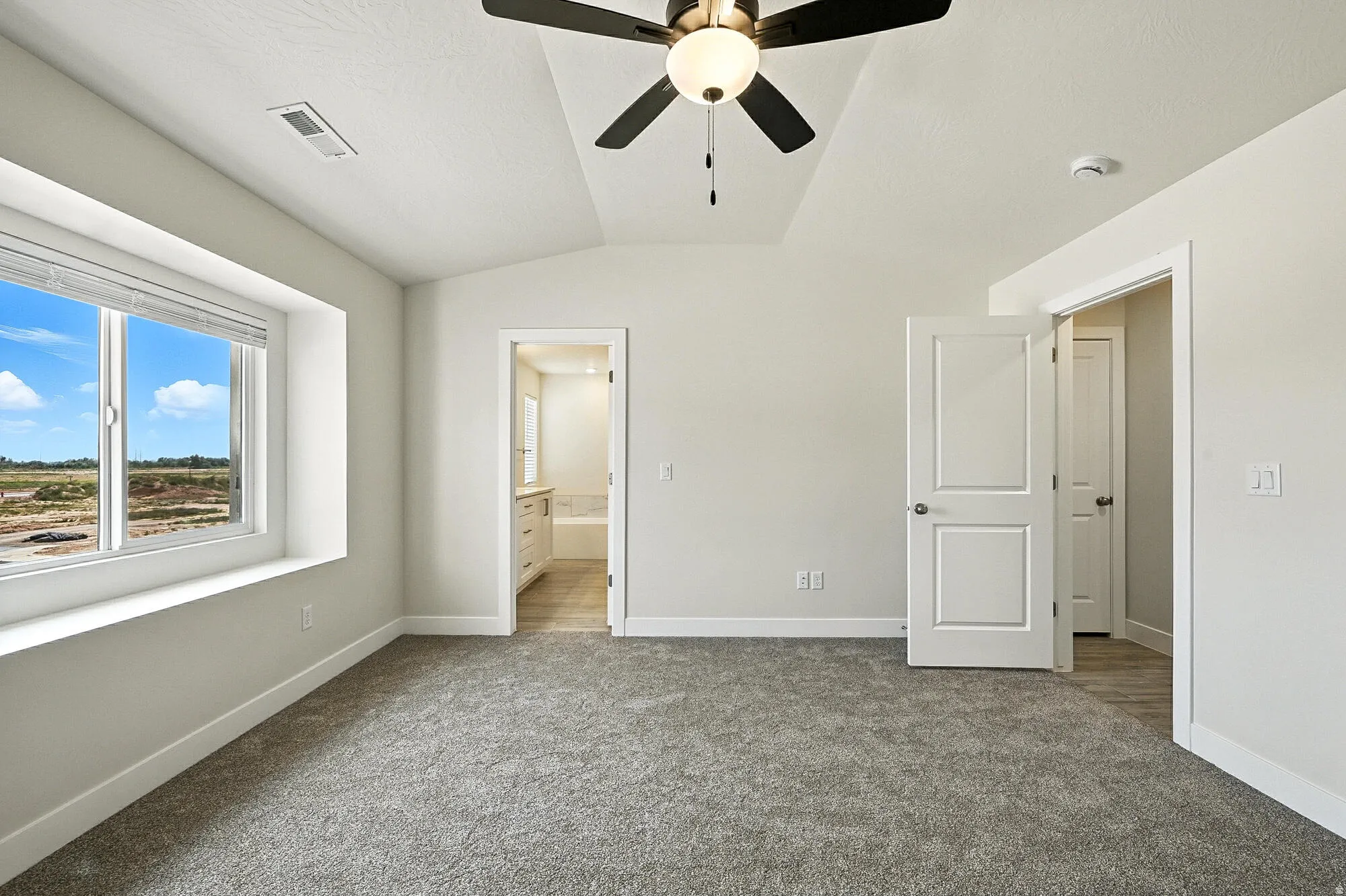 Spacious bedroom featuring vaulted ceilings, a contemporary ceiling fan with integrated lighting, plush grey carpeting, and a large window with a deep sill