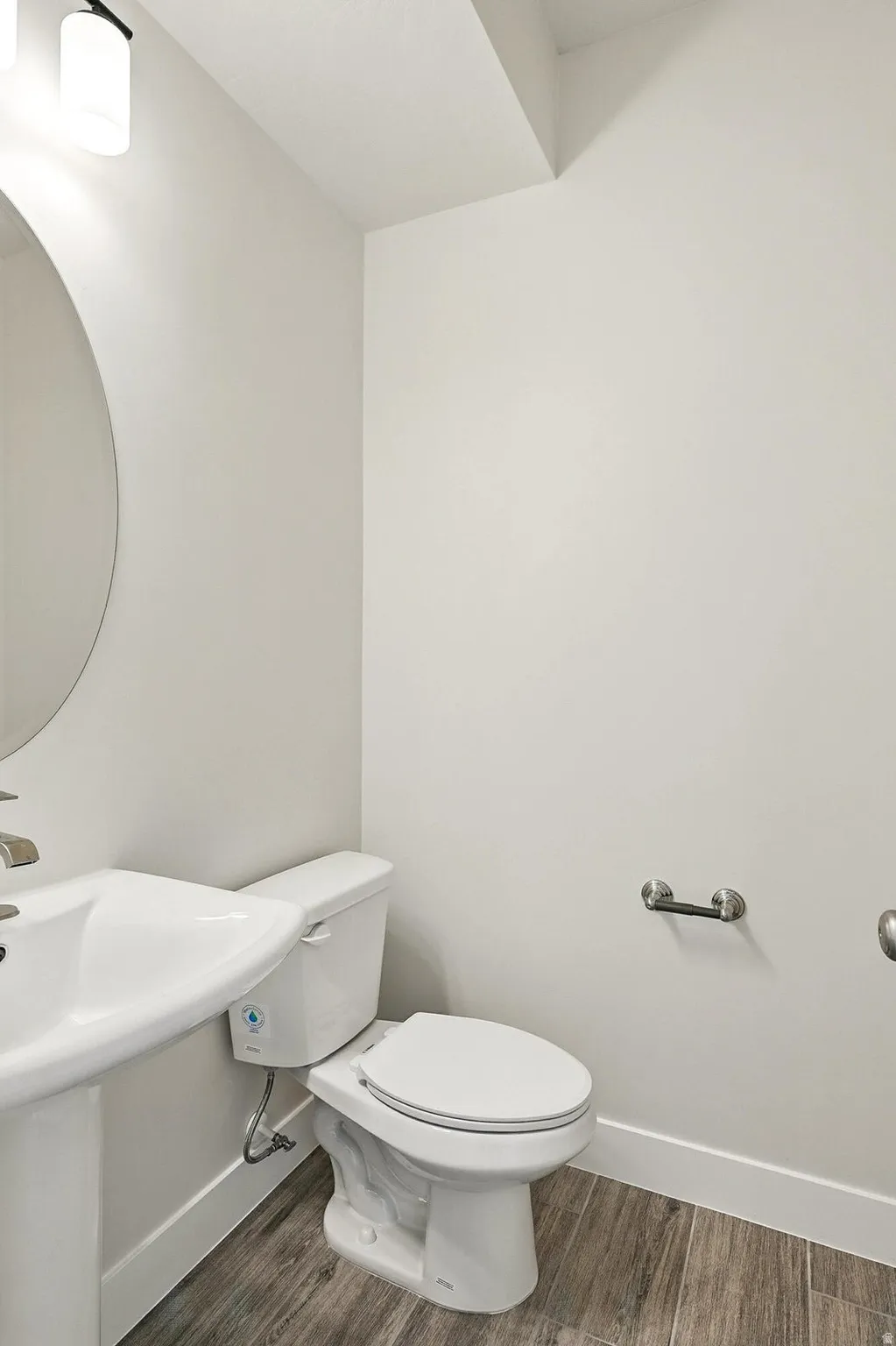 Modern powder room featuring a white pedestal sink, circular wall mirror, contemporary light fixture, and wood-finish flooring