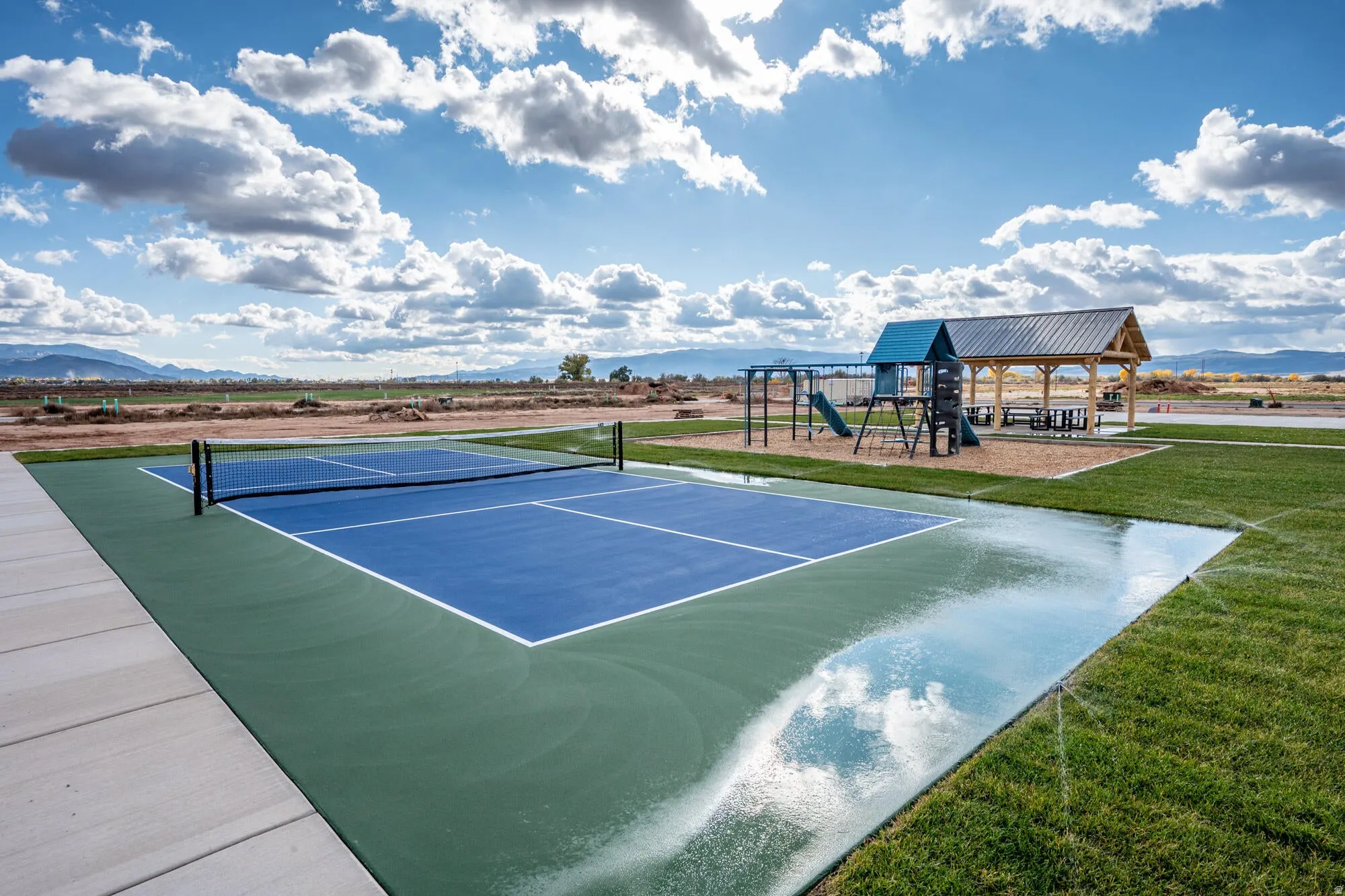 Outdoor recreational area featuring a blue and green court with a net, a playground with slides and swings, and a covered picnic area with a metal roof