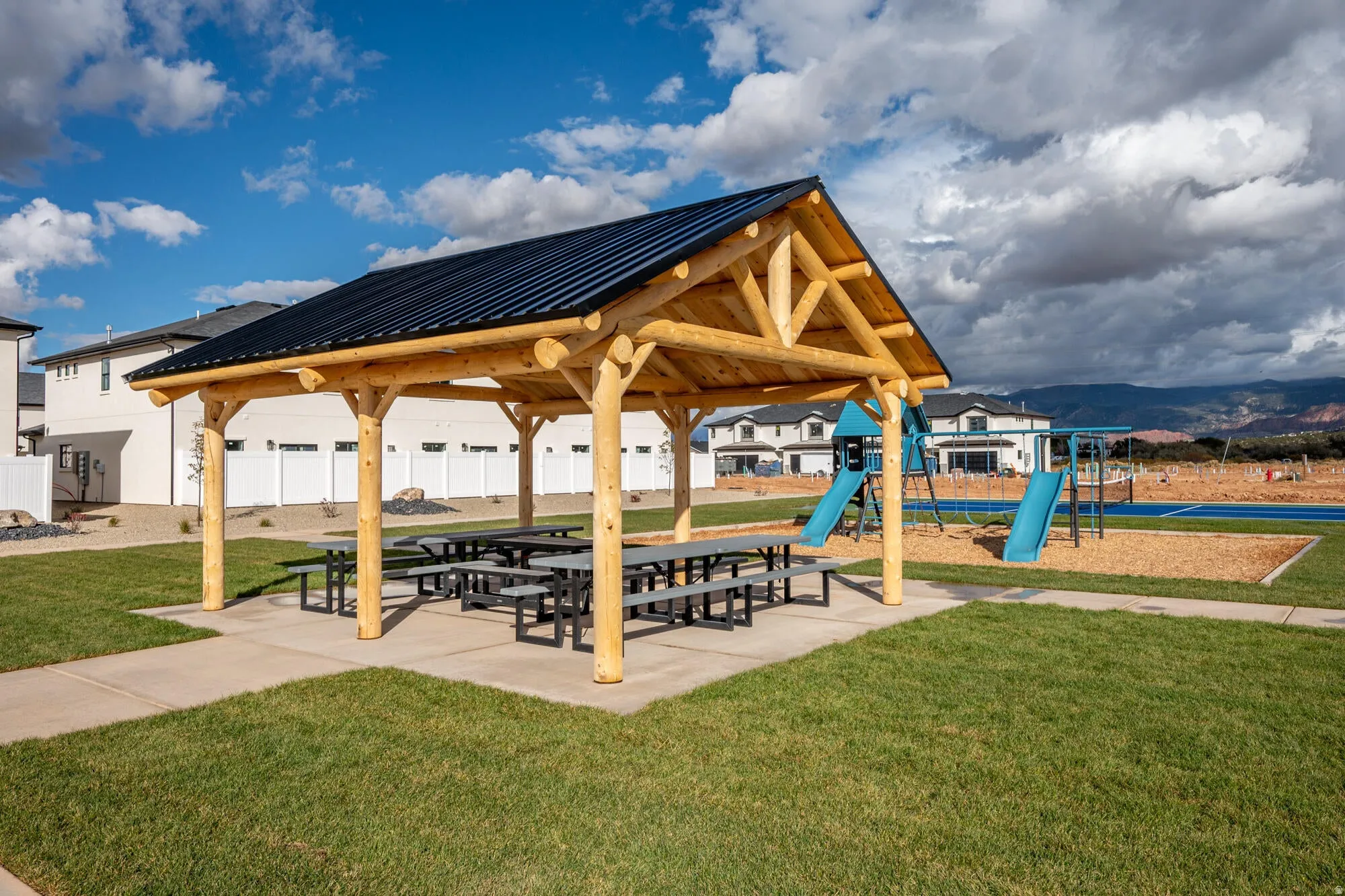 Outdoor pavilion with exposed log beam construction, dark metal roof, concrete pad, and multiple picnic tables