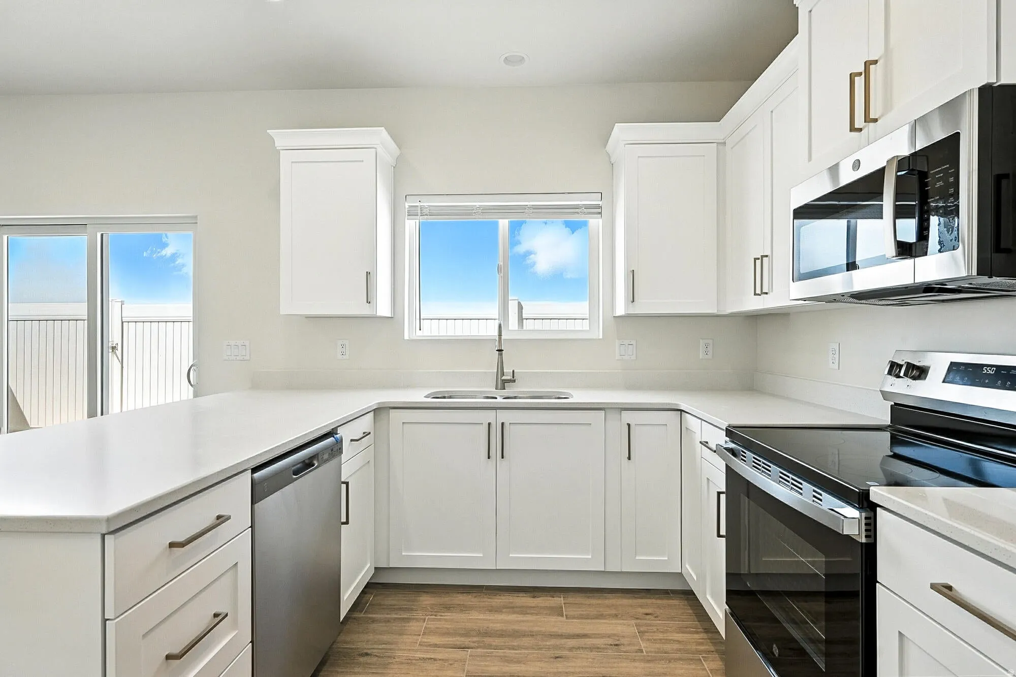 Modern kitchen featuring white shaker cabinetry, stainless steel appliances, light countertops, wood-finish flooring, and a large window above the sink
