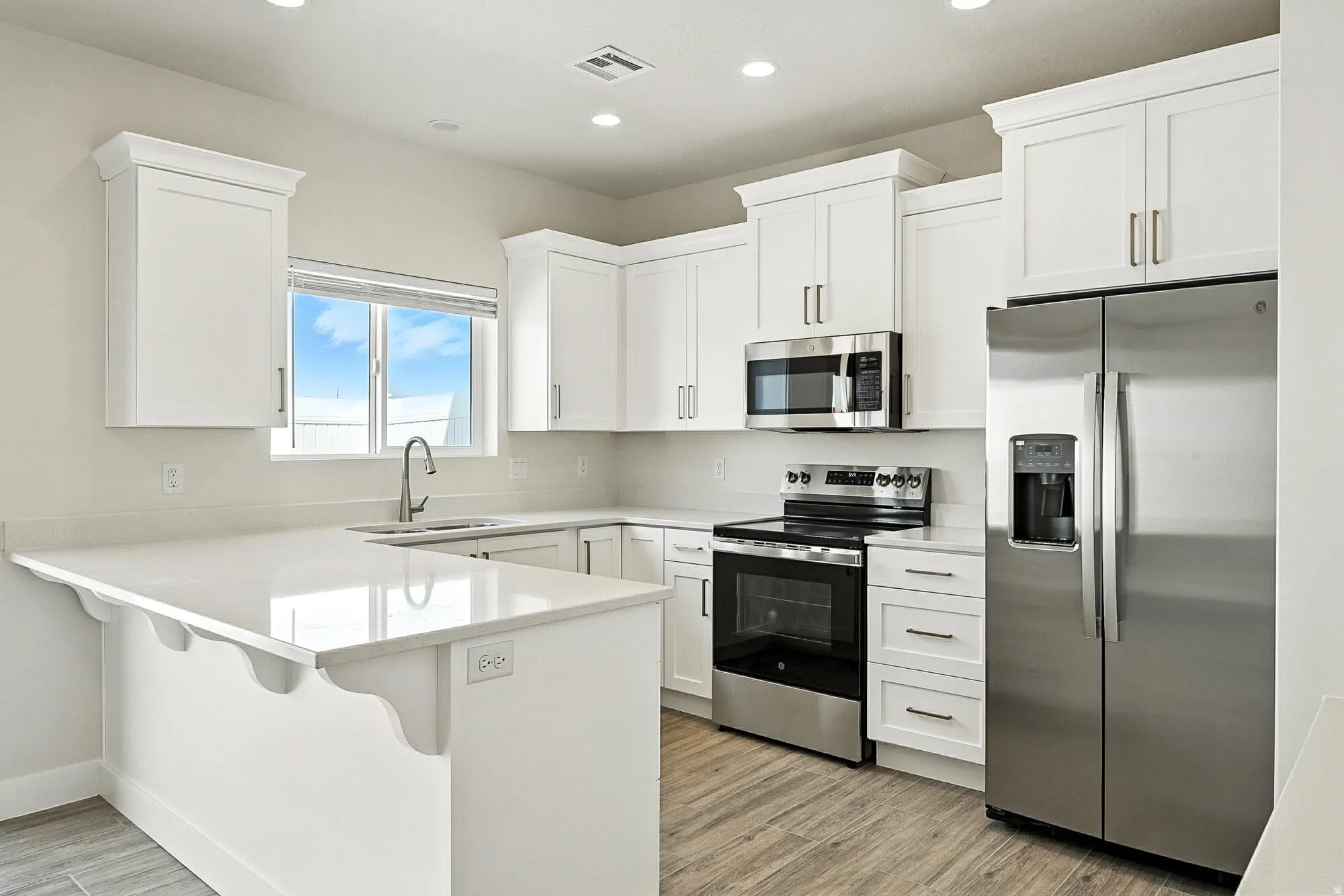 Modern kitchen featuring white shaker cabinetry, stainless steel appliances, light-toned countertops, a built-in kitchen island with decorative corbels, and wood-finish flooring