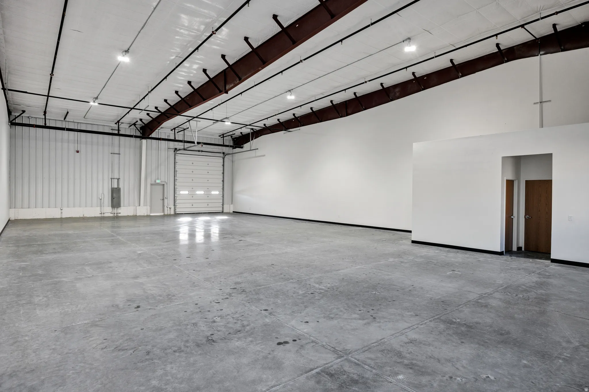 Spacious interior featuring a large roll-up door, concrete flooring, high ceilings with exposed beams, and multiple overhead light fixtures