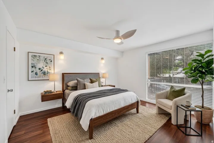 Spacious bedroom featuring dark wood-finish flooring, white walls, and a large window with horizontal blinds. Virtual Staging.
