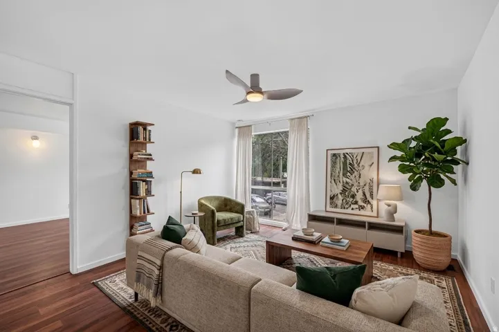 Bright living area featuring wood-finish flooring, white walls, and a contemporary ceiling fan. Virtual staging.