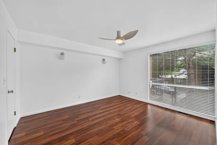 Bright room featuring rich wood-finish flooring, a contemporary ceiling fan with integrated lighting, a large window with horizontal blinds, and a white interior door