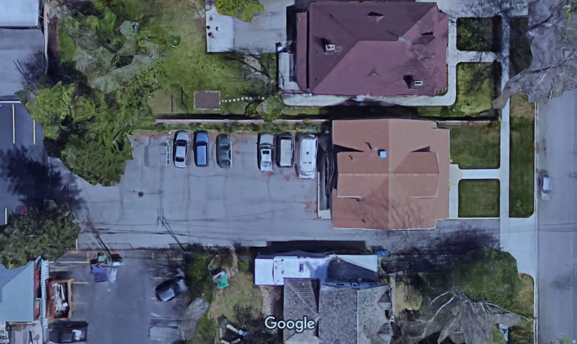 Aerial view showcasing a large paved parking area, two distinct buildings with hip roofs, and surrounding green spaces with mature trees