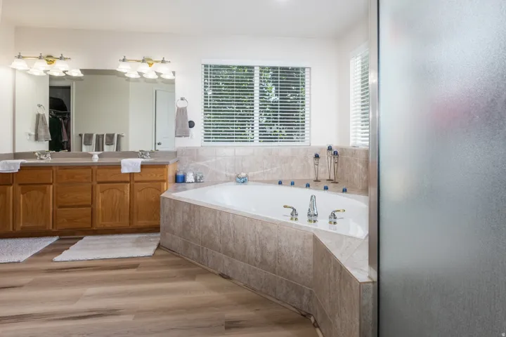 Spacious bathroom featuring a built-in corner soaking tub with tiled surround, dual vanity with wood cabinetry, large mirror, and wood-finish flooring