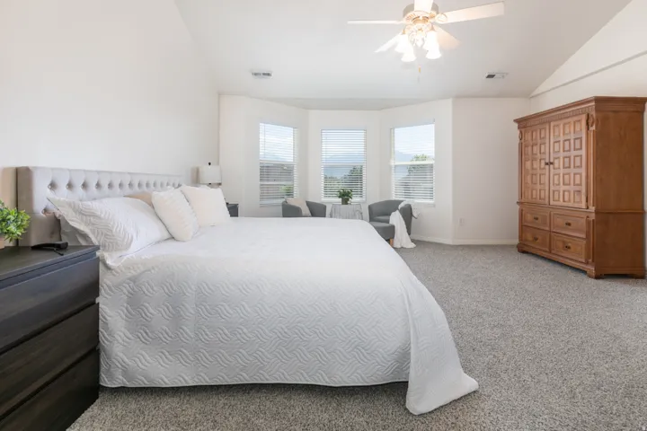 Spacious bedroom featuring plush carpeting, a large bay window with horizontal blinds, and a vaulted ceiling with a ceiling fan