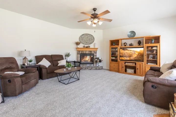 Living area featuring light-colored carpeting, a stone-faced fireplace with a wood mantle, a built-in entertainment center with display cabinets, a ceiling fan with light fixture, and neutral wall paint