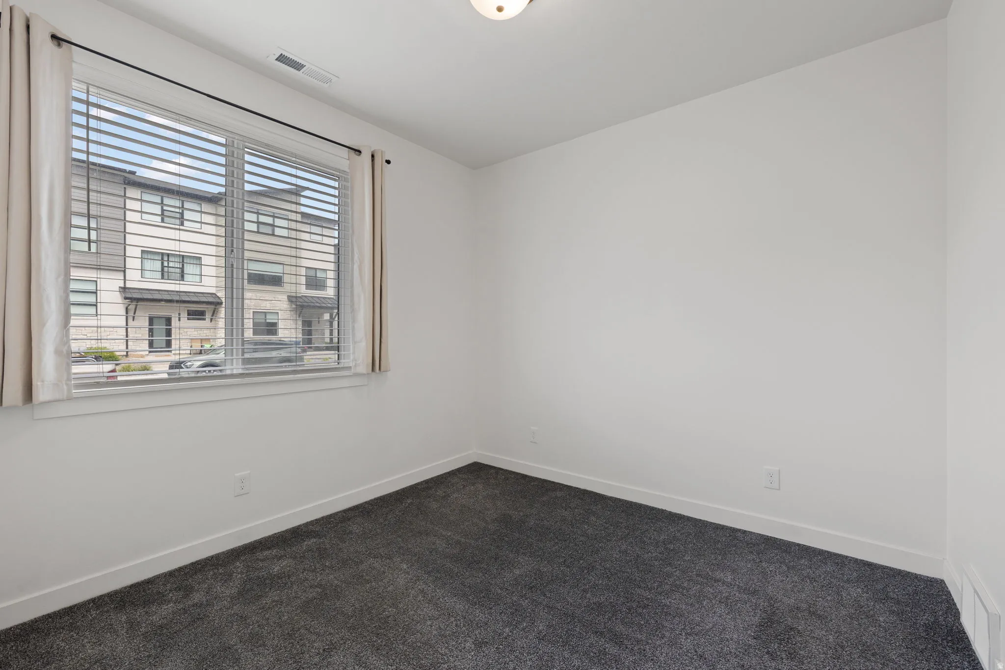 Bright interior space featuring a large window with blinds and curtains, neutral wall paint, and dark gray carpeting