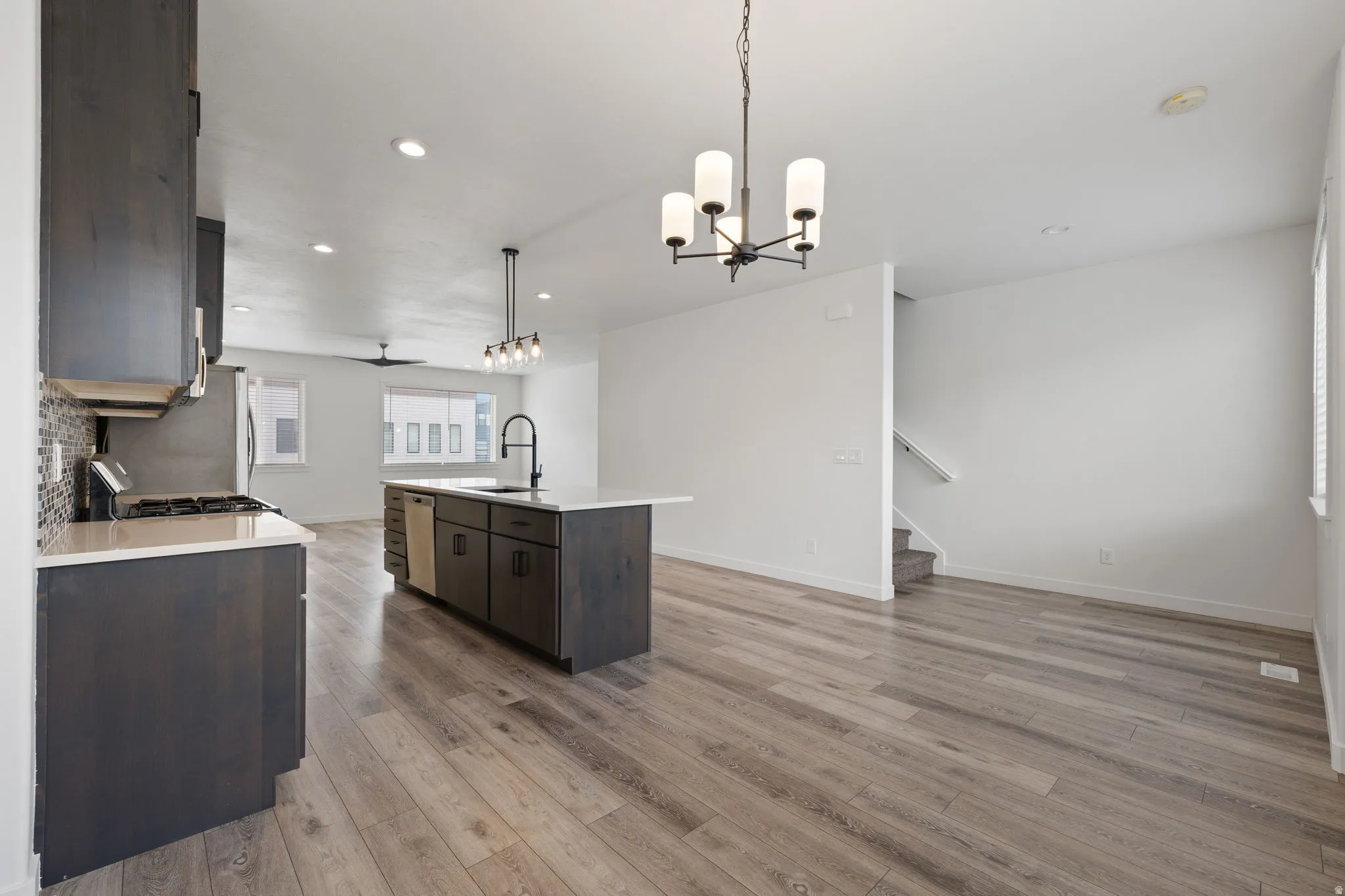 Open-concept living space featuring wood-finish flooring, a kitchen island with integrated sink, dark wood cabinetry, and a tile backsplash