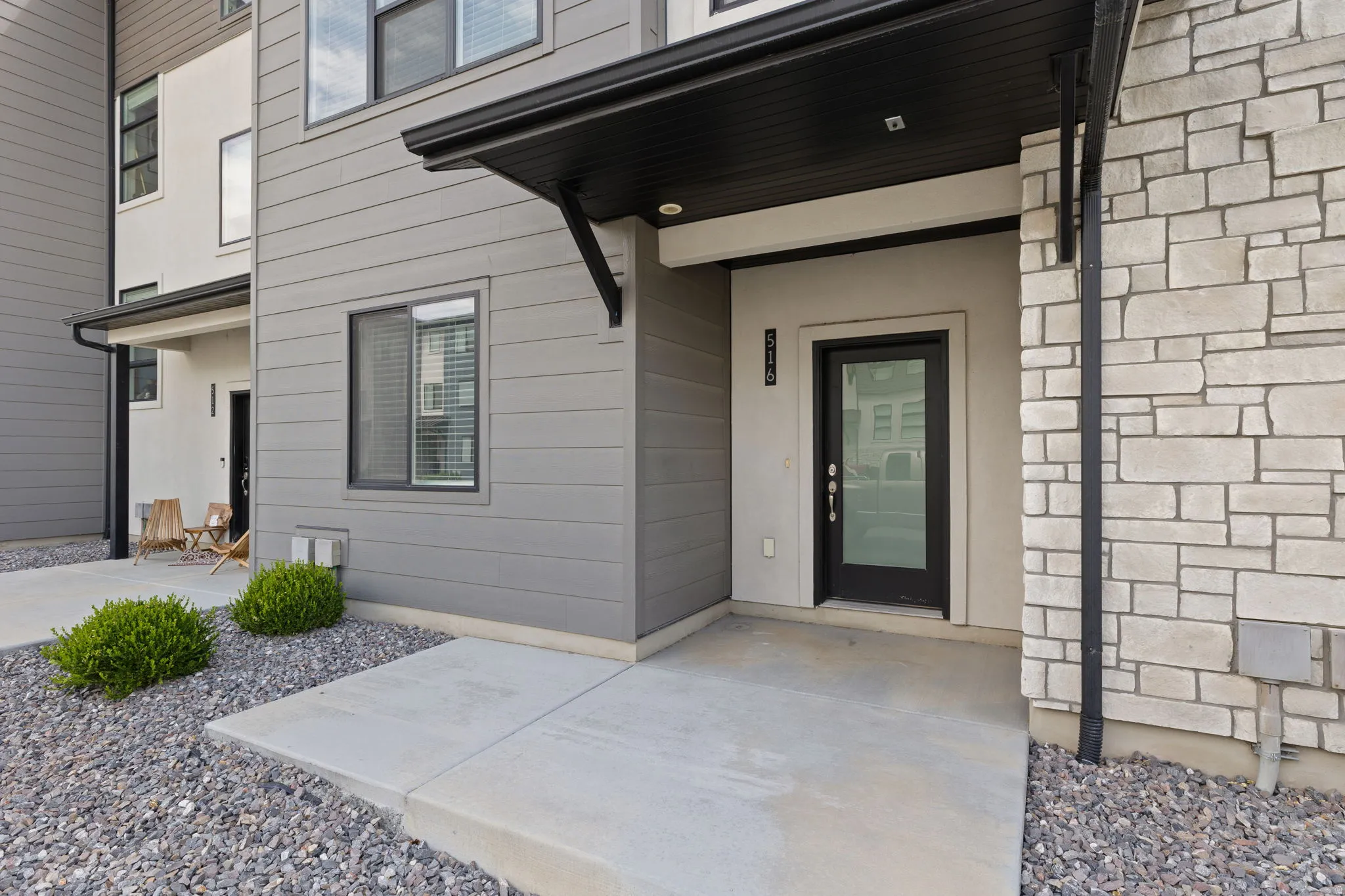 Modern exterior featuring a prominent black framed glass entry door, light stone facade, horizontal gray siding, a covered entry, and concrete pathways with gravel landscaping