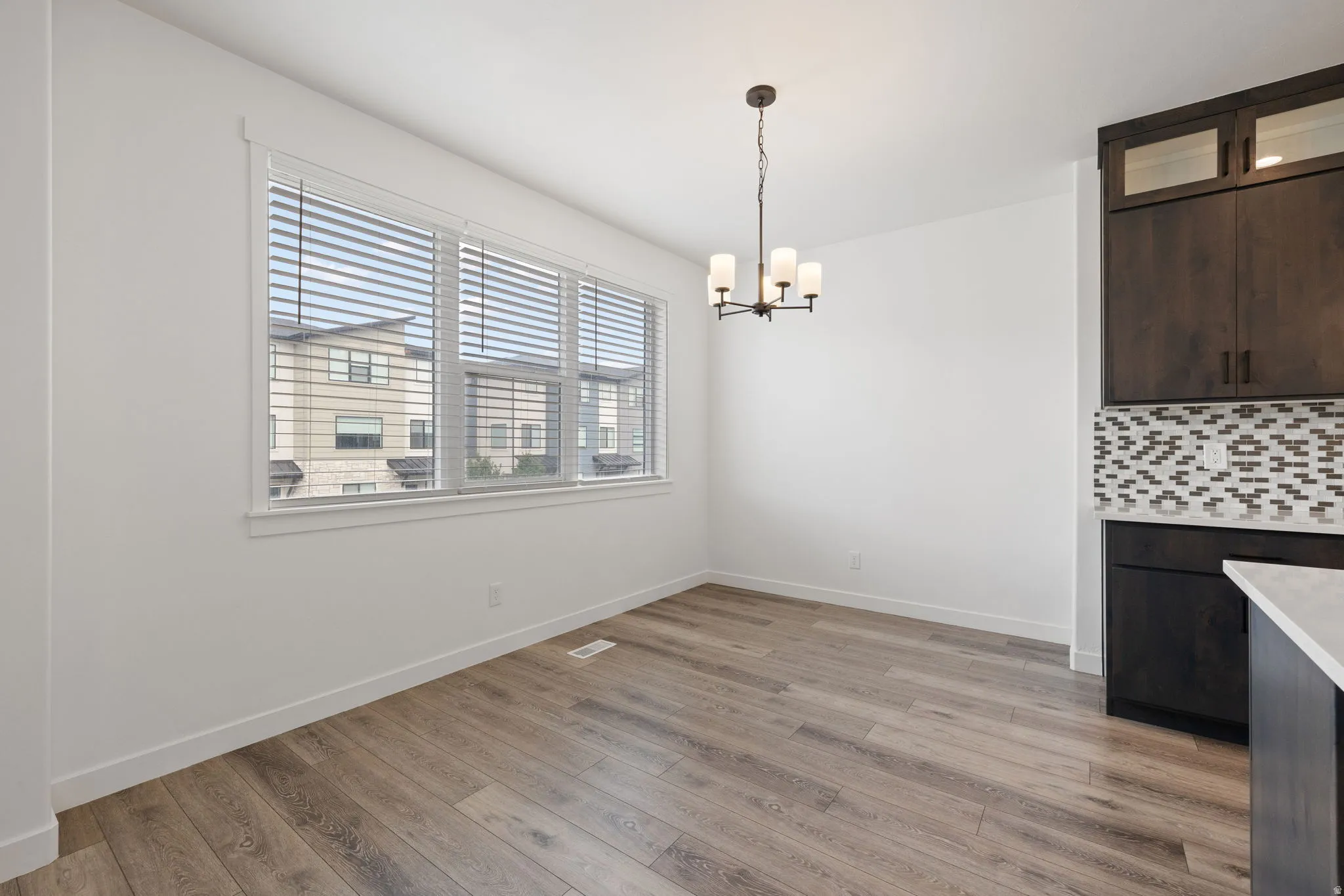 Spacious dining area featuring wood-finish flooring, ample natural light from large windows, white baseboards, a contemporary chandelier, and a built-in cabinetry with a patterned tile backsplash