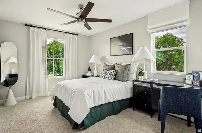 Neutral-toned room featuring a ceiling fan with wood-finish blades, two windows with white trim, and light-colored carpet