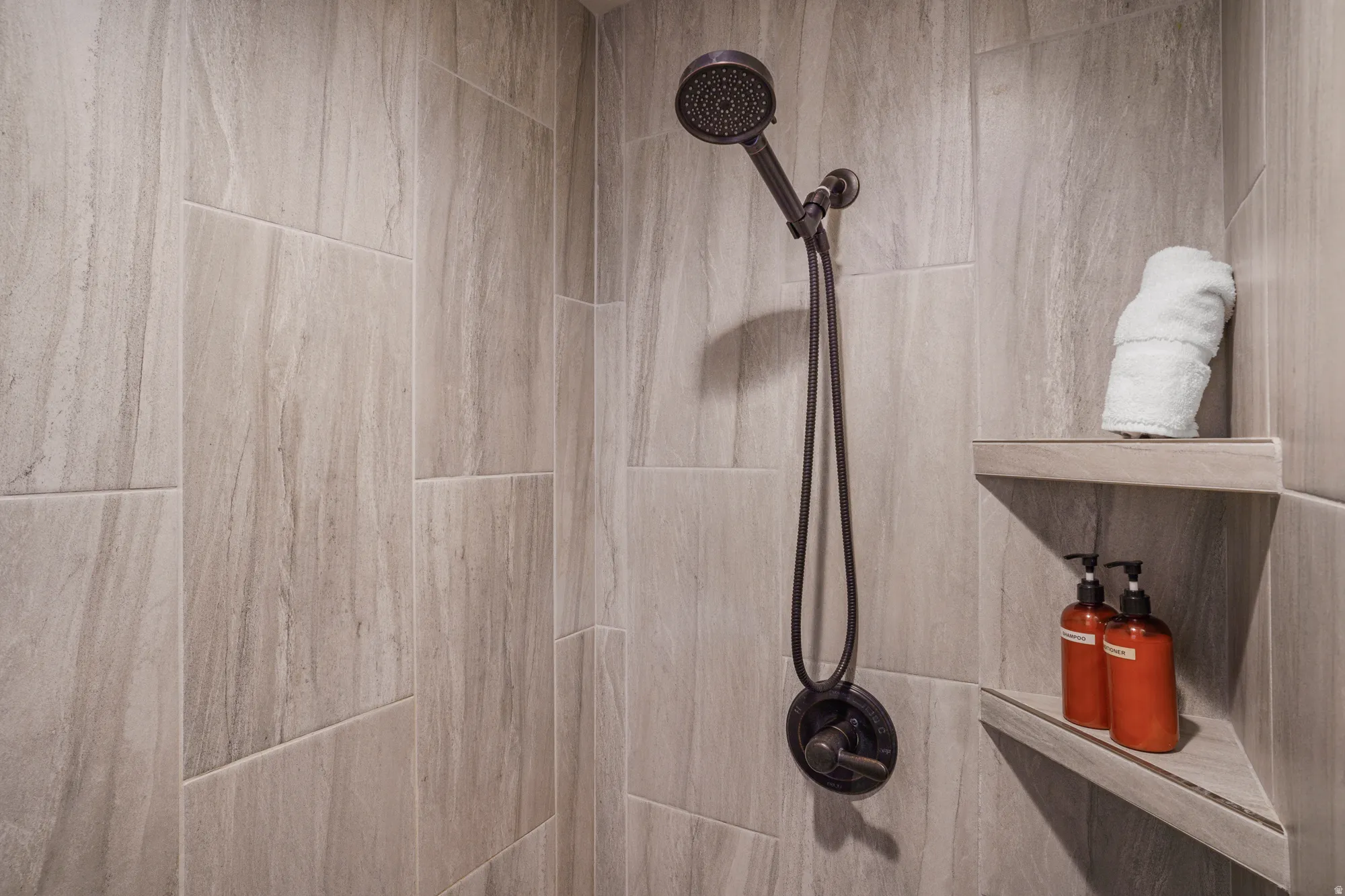 Shower with large format stone-look tile, dark bronze showerhead, and integrated corner shelving