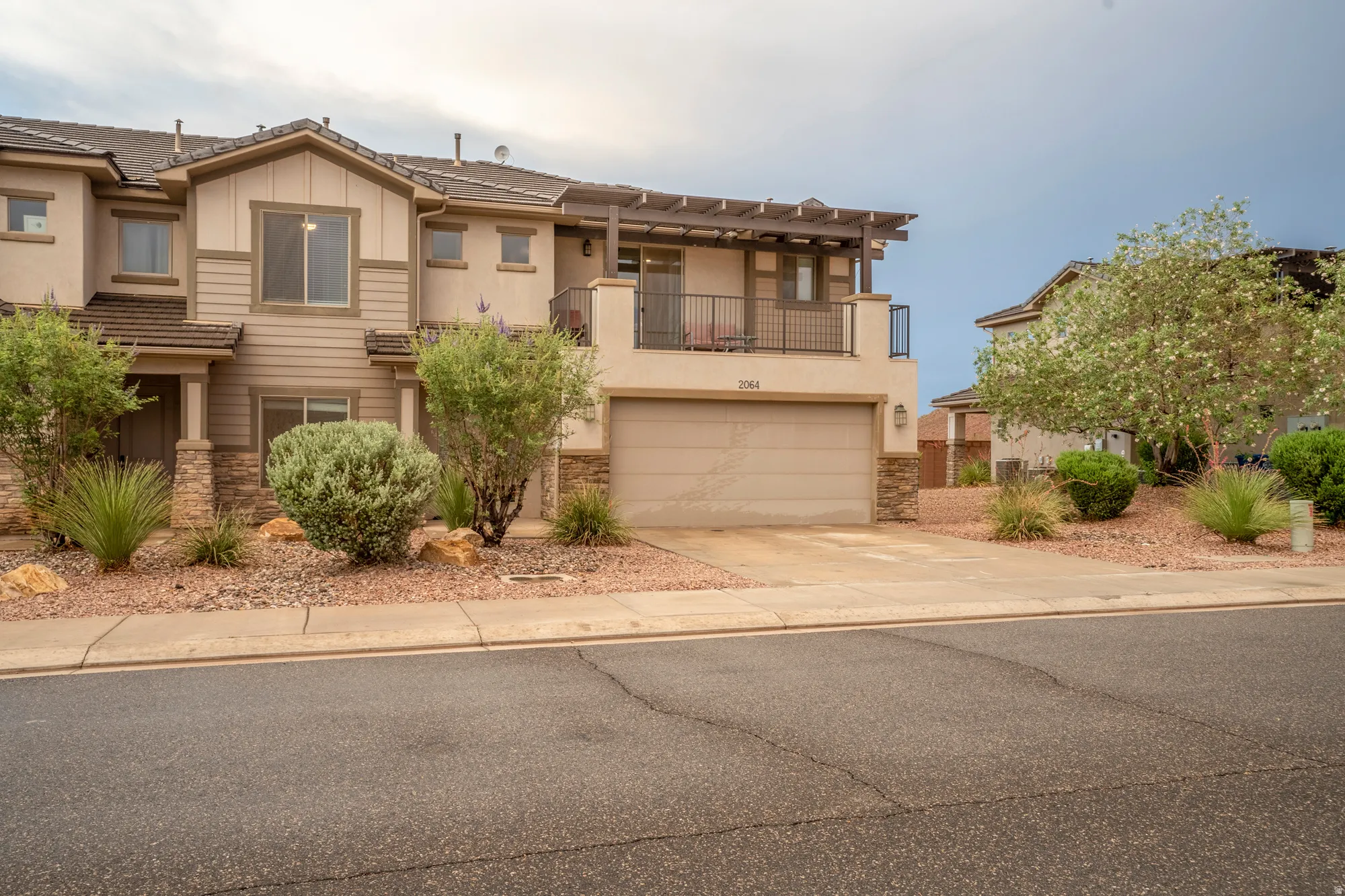 Contemporary townhome with a stucco and stone facade, featuring a two-car garage, upper-level balcony, and xeriscaped front yard with mature shrubbery and rock accents