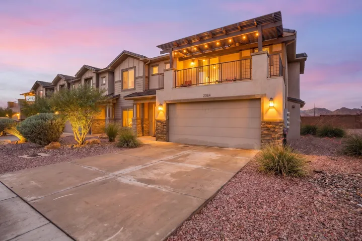 Contemporary multi-level residence featuring a stucco and stone facade, an integrated garage, a second-story balcony with an open pergola, and desert landscaping with accent lighting