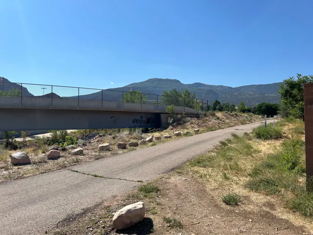 Paved pathway bordered by natural rock formations, with a concrete and chain-link fence bridge spanning a dry creek bed