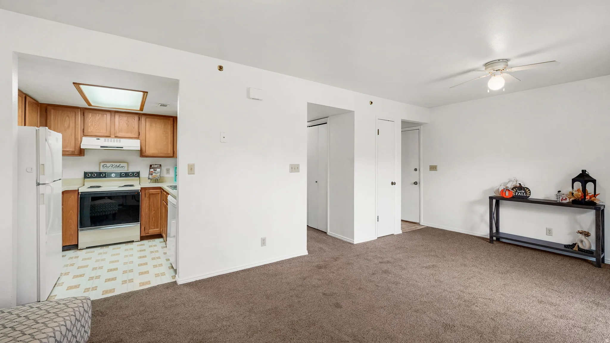 Living area with carpeting and ceiling fan, featuring a kitchen entry with wood-finish cabinetry, white countertops, built-in range, and a white refrigerator