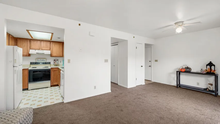 Living area with carpeting and ceiling fan, featuring a kitchen entry with wood-finish cabinetry, white countertops, built-in range, and a white refrigerator