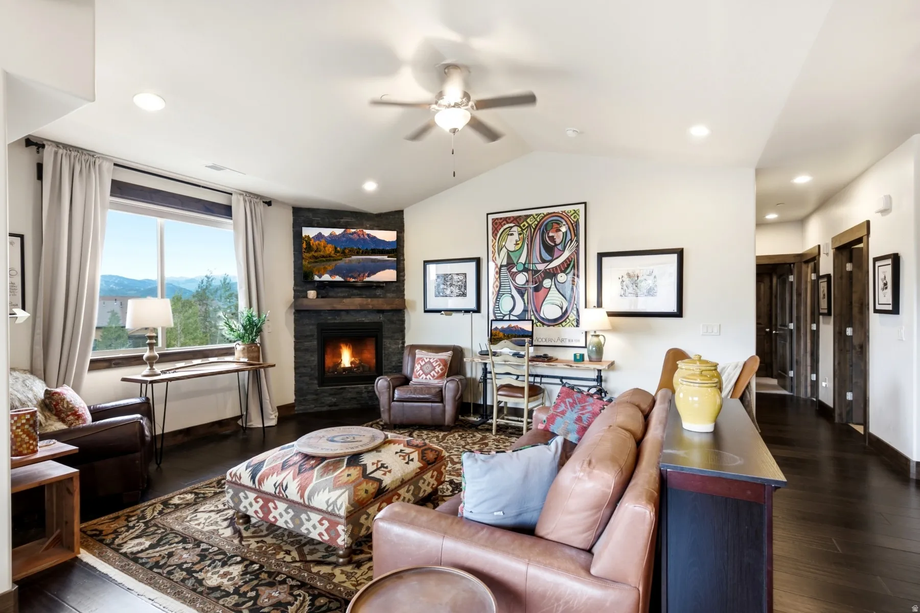 Spacious living area featuring dark wood-finish flooring, a corner fireplace with stone surround, a large window offering expansive mountain scenery, vaulted ceilings, and recessed lighting