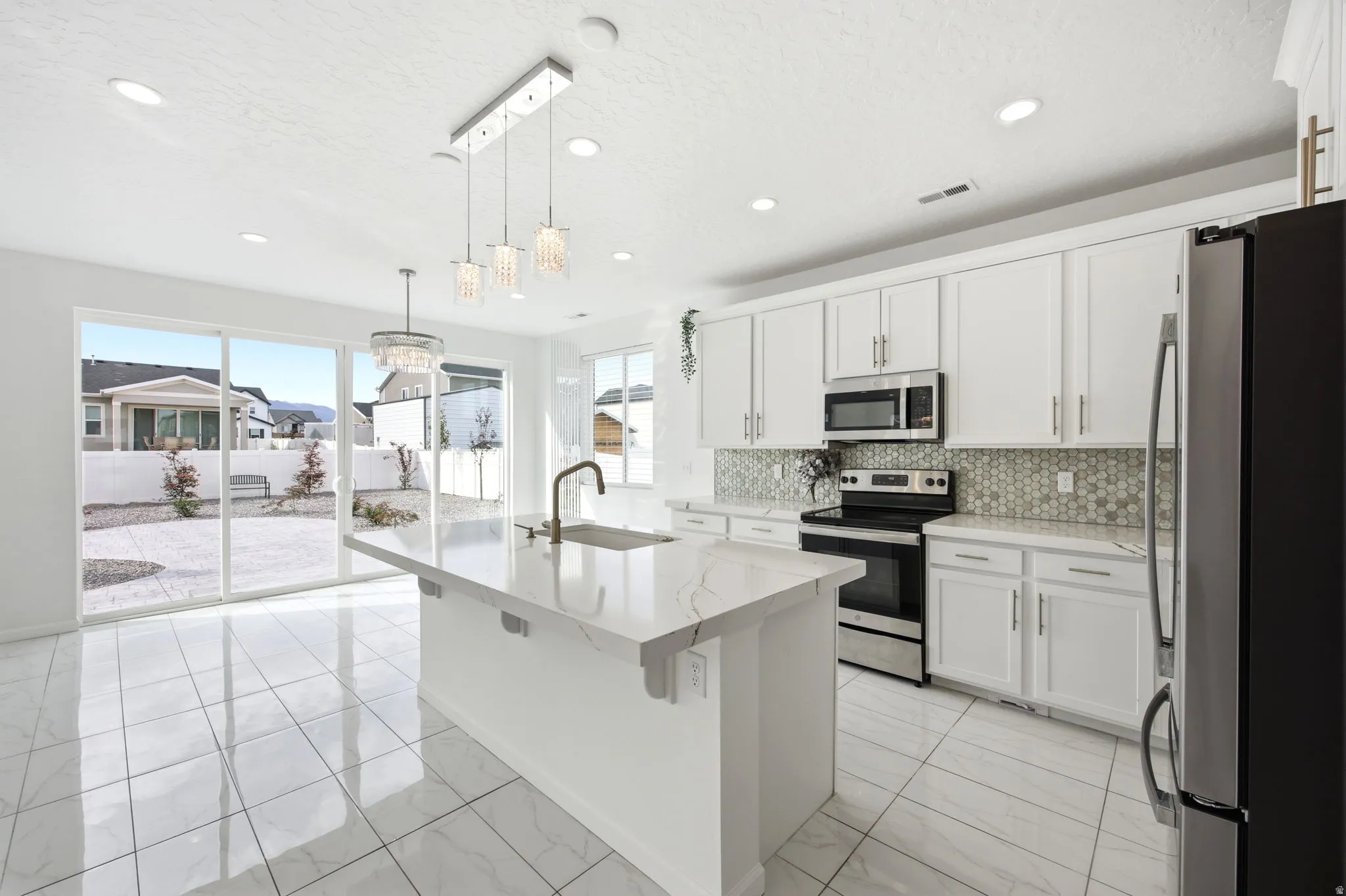 Open-concept kitchen featuring an island with a sink and modern pendant lighting, white cabinetry, integrated stainless-steel appliances, and a mosaic tile backsplash