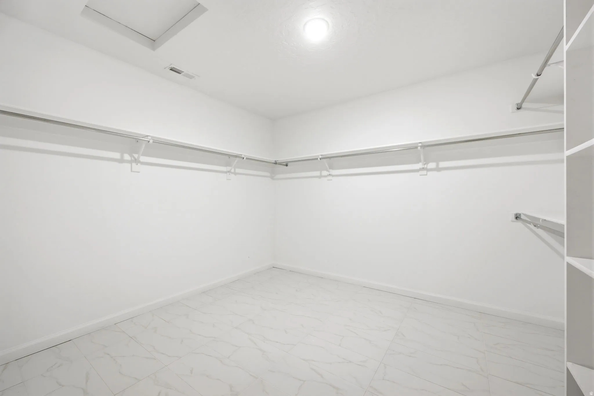 Spacious closet featuring white walls, white-finish shelving, metal hanging rods, and large format white tile flooring