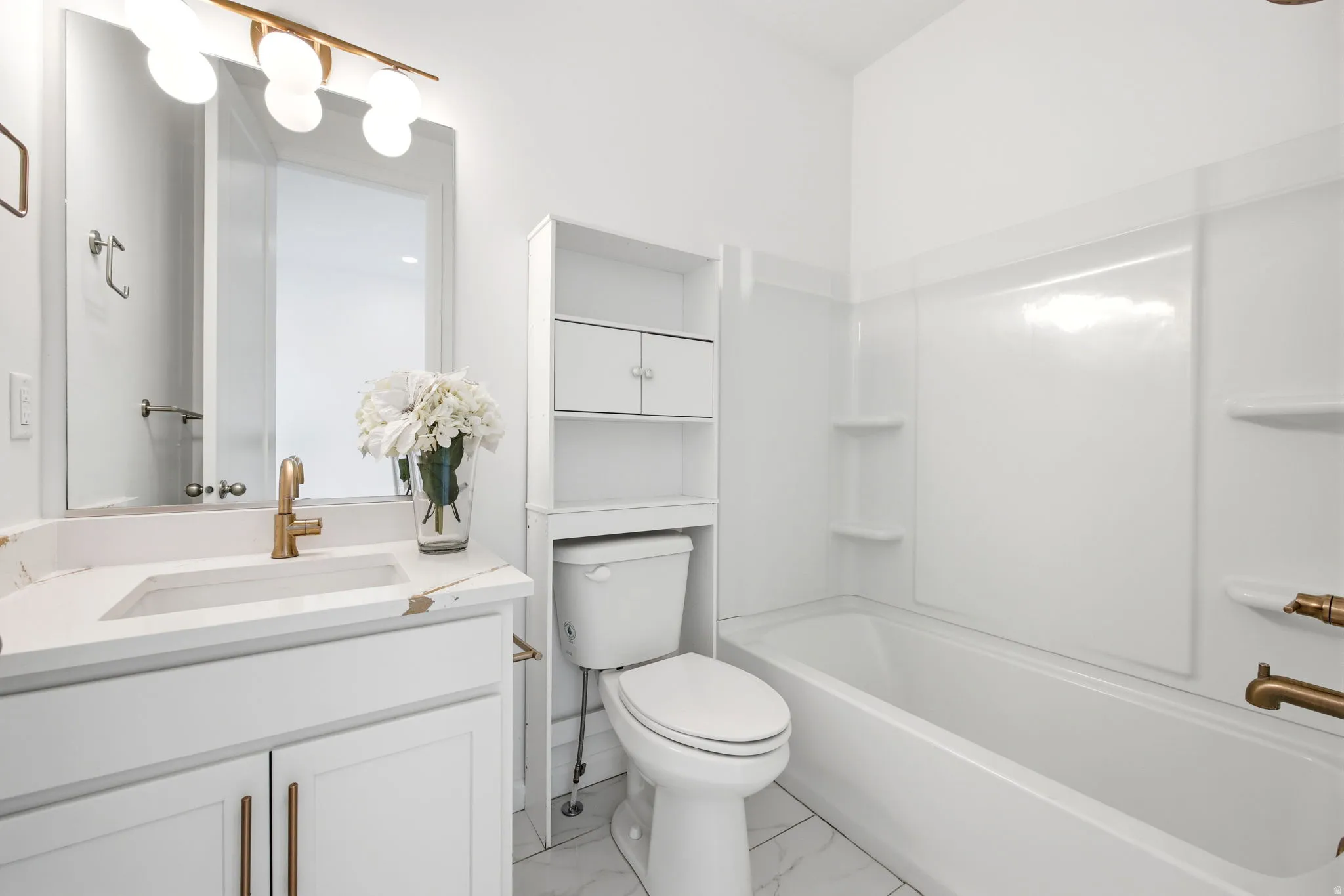 Full bathroom featuring a white vanity with a gold-tone faucet, a mirror with a spherical light fixture, a toilet with an over-the-toilet storage unit, a built-in bathtub with a white surround, and white tile flooring