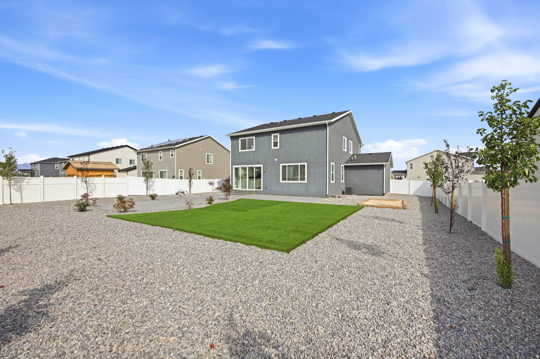 Expansive backyard featuring a central artificial turf area, surrounding gravel landscaping, a white privacy fence, and scattered young trees