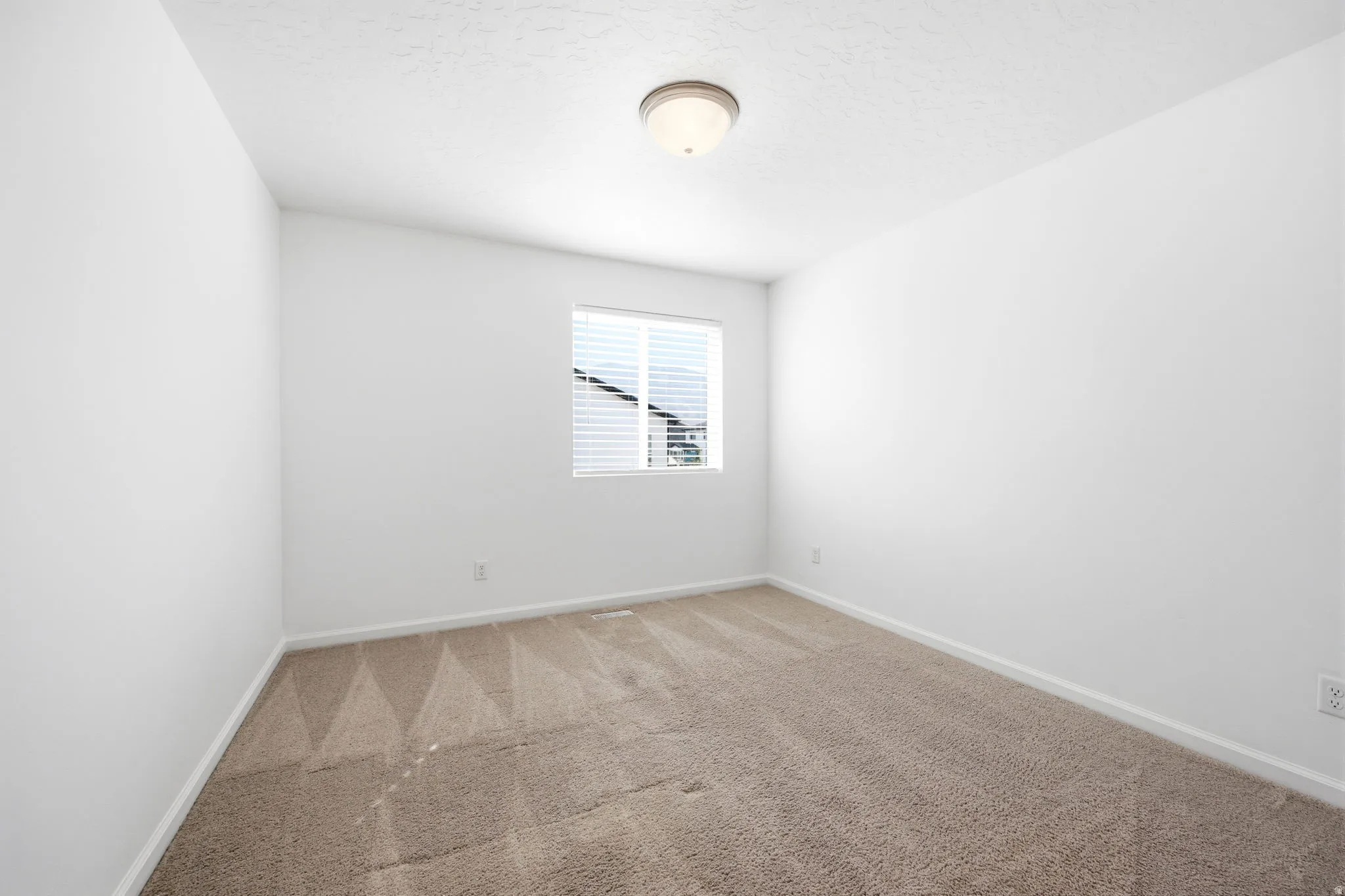 Neutral tone-on-tone carpet flooring, white walls, and a single window with horizontal blinds provide a versatile interior space