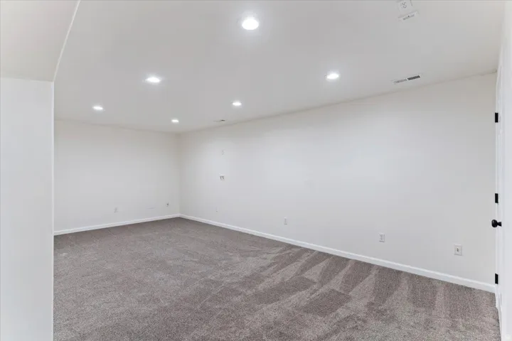 Spacious room featuring neutral carpeting, recessed ceiling lighting, crisp white walls, white baseboards, and a white interior door with matte black hardware