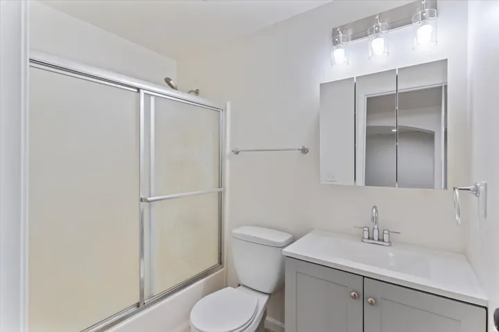 Bathroom featuring a white bathtub with frosted sliding shower doors, a commode, and a vanity with a white countertop and gray cabinetry