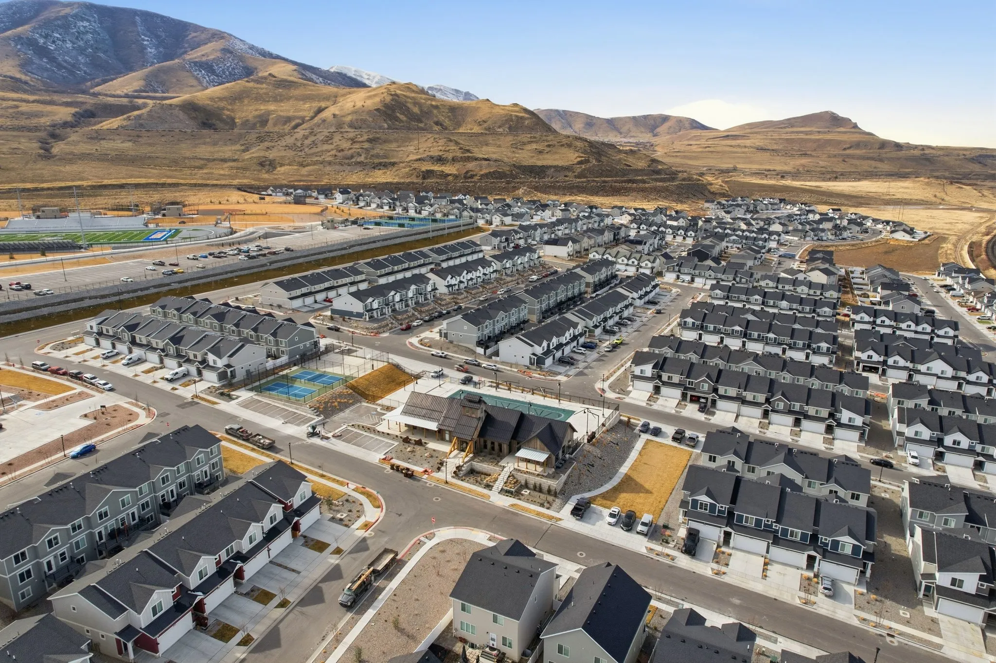 Expansive community featuring numerous residential structures with dark roofs and light siding, a central clubhouse, and multiple sports courts