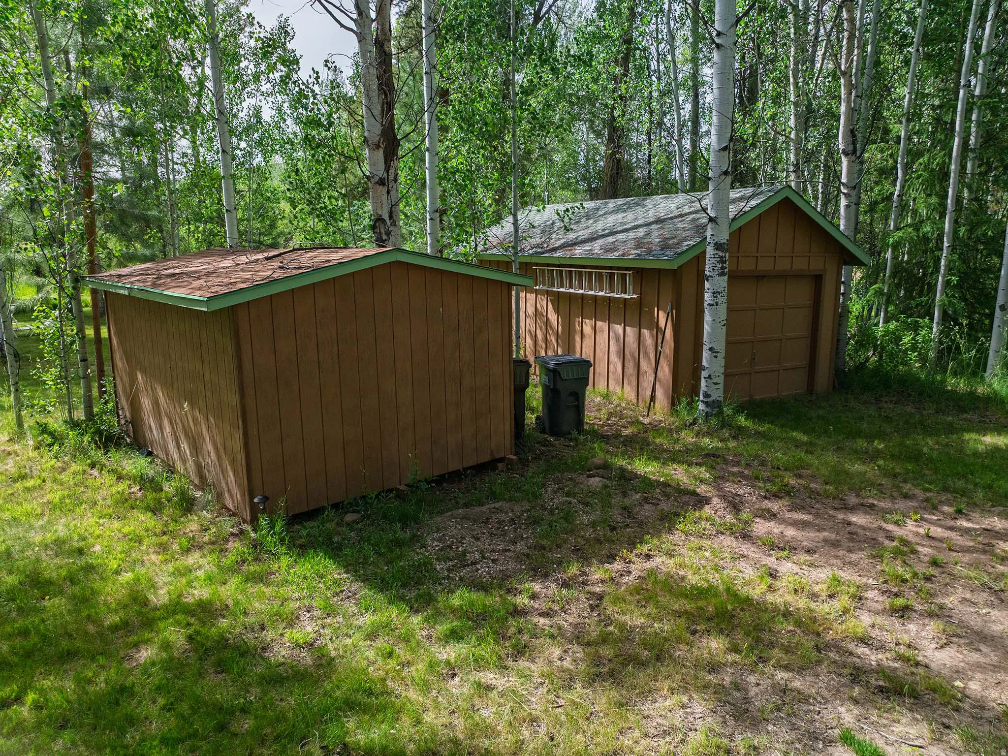 Two brown wood-sided outbuildings featuring shingle roofs and green trim, surrounded by mature trees