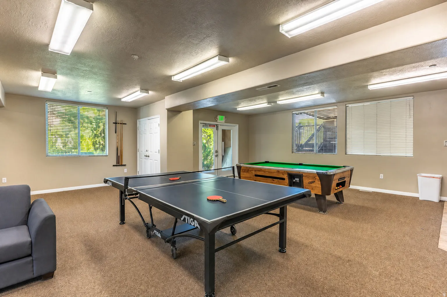 Spacious recreation room featuring carpet flooring, multiple windows with blinds, and overhead fluorescent lighting
