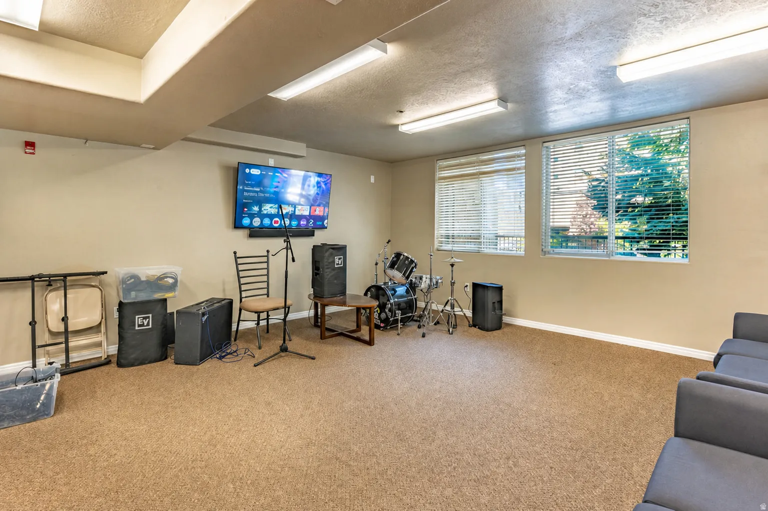Spacious room featuring carpeting, two windows with blinds, a wall-mounted television, and overhead fluorescent lighting