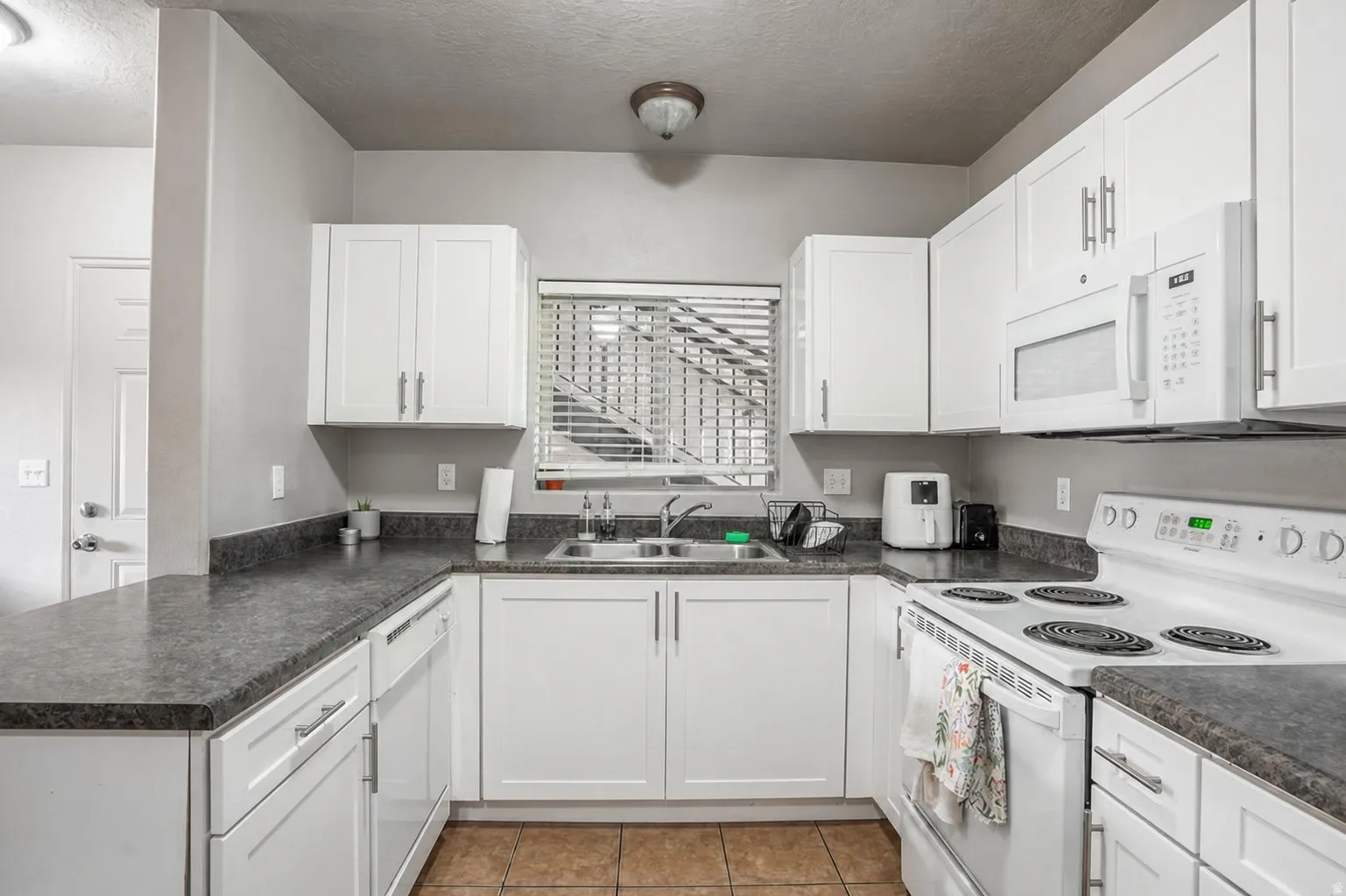Kitchen featuring white cabinetry, dark countertops, a double basin stainless steel sink, tile flooring, and white appliances including a range and built-in microwave