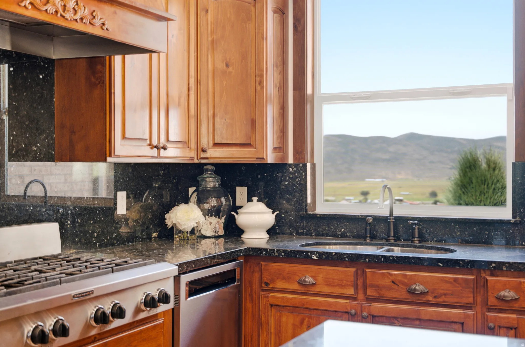 Kitchen featuring custom wood cabinetry, speckled dark countertops with matching backsplash, stainless steel gas range, and a window overlooking distant hills