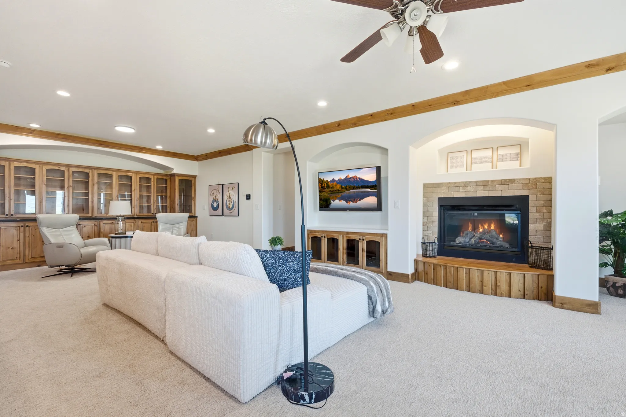 Recessed lighting illuminates a carpeted living space featuring a built-in fireplace with a stone surround and wood-finish hearth, a ceiling fan, and built-in cabinetry with glass doors