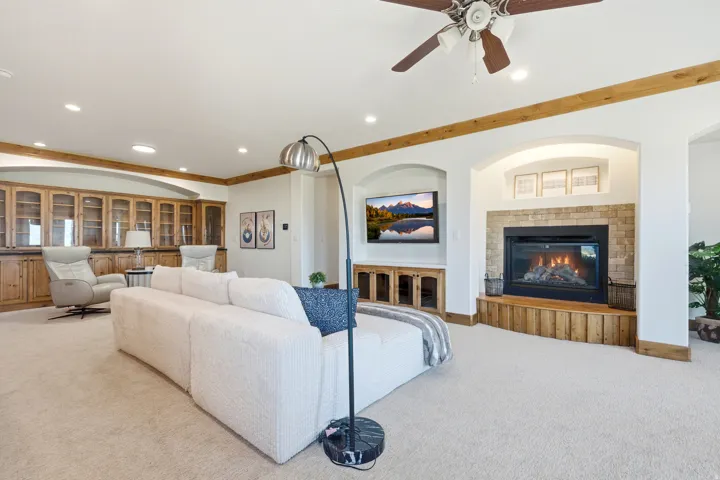 Recessed lighting illuminates a carpeted living space featuring a built-in fireplace with a stone surround and wood-finish hearth, a ceiling fan, and built-in cabinetry with glass doors