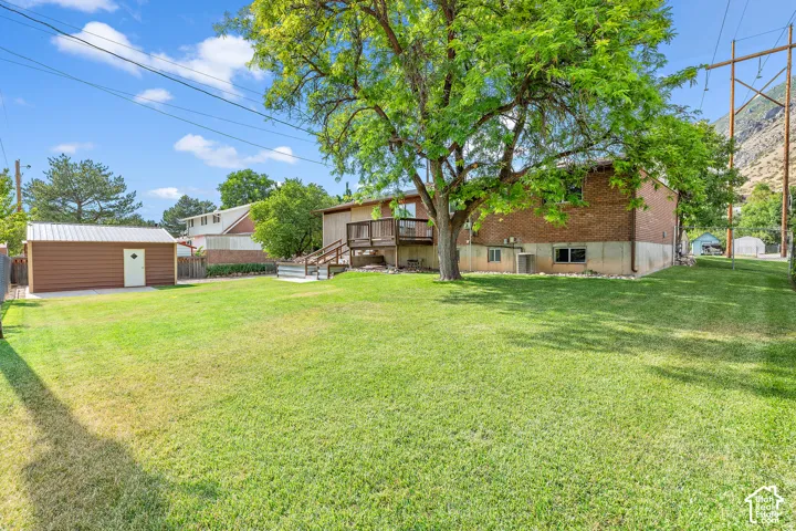 Expansive grassy yard featuring a large shade tree, a brick-sided residence with a raised deck, a separate brown outbuilding with a white door, and a mountainous backdrop