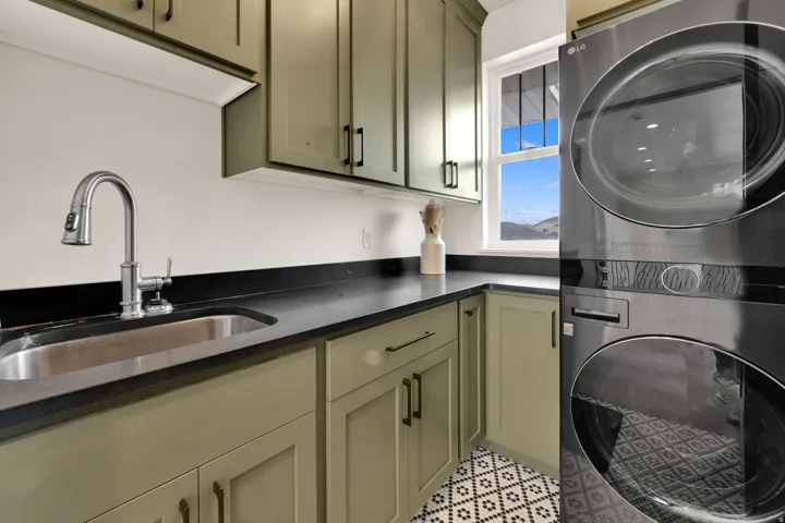 Utility space with stacked washer and dryer, olive green cabinetry, dark countertops, and a stainless steel sink