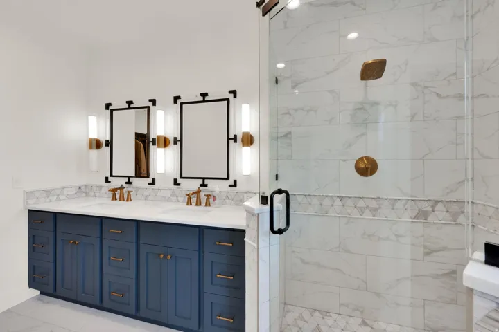 Spacious bathroom featuring a dual vanity with a blue cabinet base and stone countertop, dual framed mirrors with vertical sconce lighting, and a glass-enclosed shower with gold-finish fixtures and decorative tilework