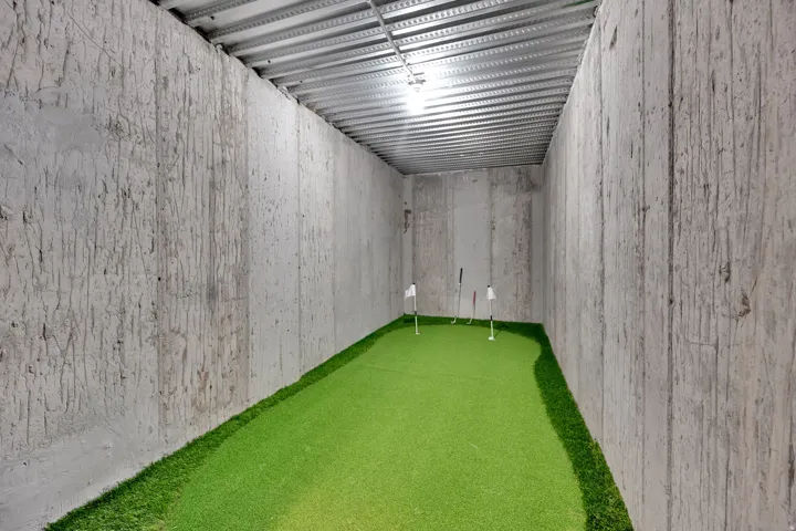 Indoor putting green with multiple holes, artificial turf, board-formed concrete walls, and a metal ceiling
