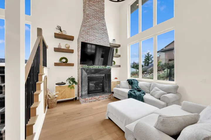 Vaulted living room featuring a floor-to-ceiling brick fireplace with a stone mantel, light wood-finish flooring, a contemporary staircase with black spindles and light wood handrails, and large vertical windows extending to the ceiling