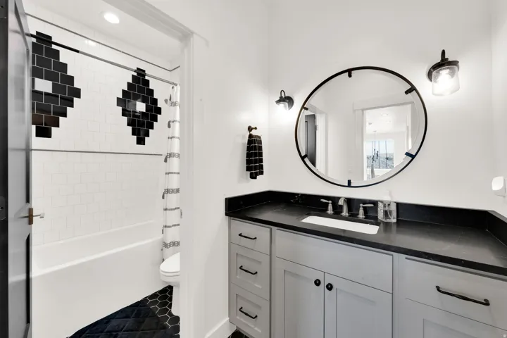 Bathroom featuring a white subway tile tub surround with black accent tiles, a dark hexagon tile floor, a light gray vanity with dark hardware, a dark countertop, and a round wall mirror